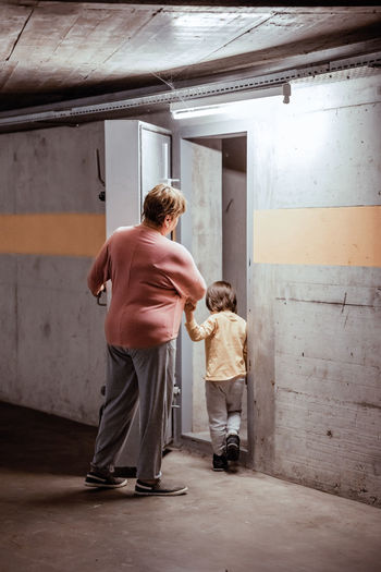 Senior woman with little child entering nuclear fallout shelter built in basement of building 