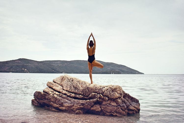 Rear view of woman in swimsuit doing yoga on rock by sea against sky