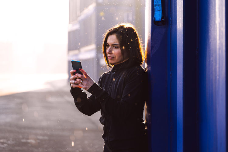 Young adult female runner is using smartphone among the shipping containers of a commercial port