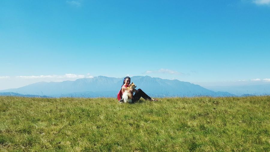 Woman sitting with dog on grassy field against blue sky
