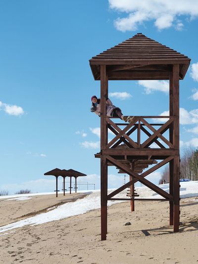 Woman sitting on railing of lifeguard hut at beach against sky