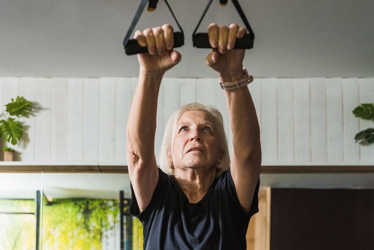 Focused elderly female athlete in sportswear with gray hair working out with straps while in the gym