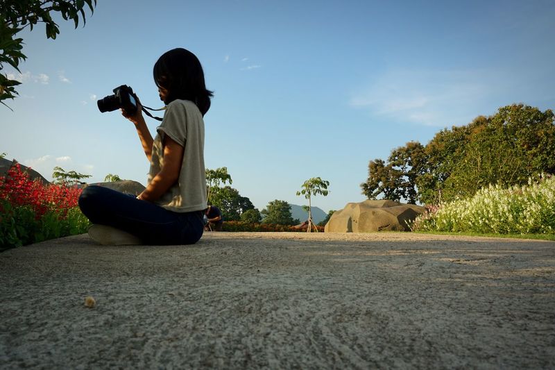 Side view of woman photographing while sitting on footpath