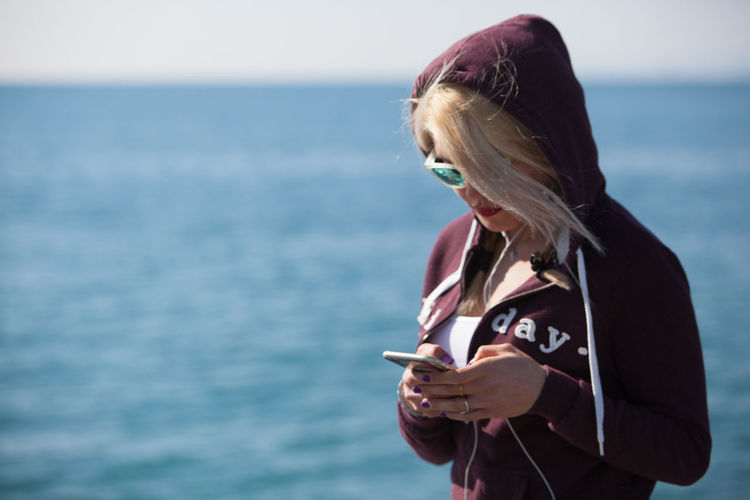 Woman in hooded shirt using mobile phone against sea