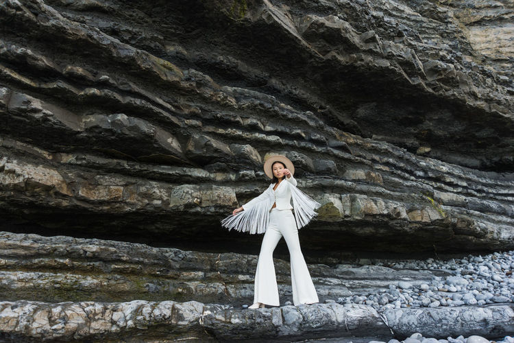Full body of elegant young slim female model in white suit with wings shaped tassel sleeves and hat outstretching arms while standing against rough rocky formation