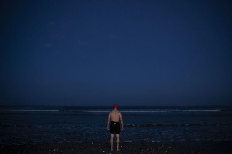 Man in swimming shorts standing at water's edge during dawn