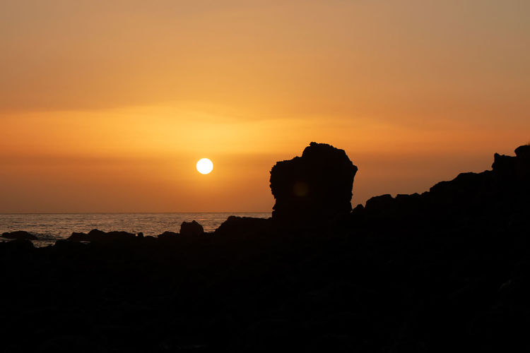 Silhouette rock formation by sea against orange sky