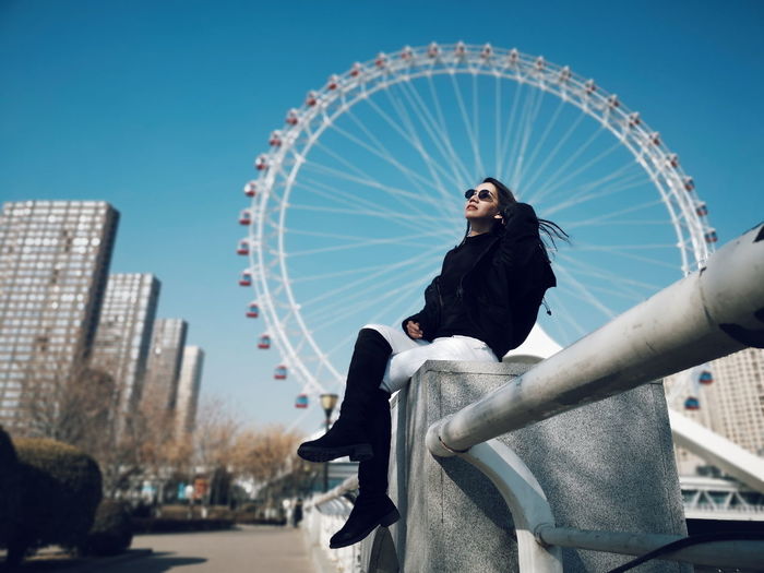 Low angle view of woman sitting on retaining wall against ferris wheel and sky