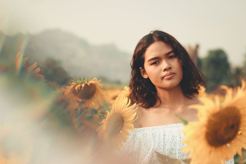 Portrait of young woman standing at sunflower farm against clear sky