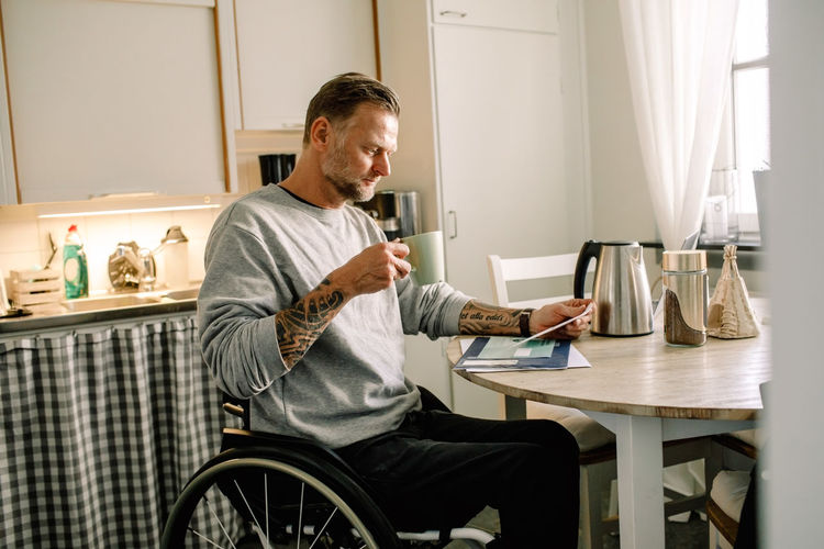 Tattooed mature man reading mail while holding coffee cup on wheelchair at home