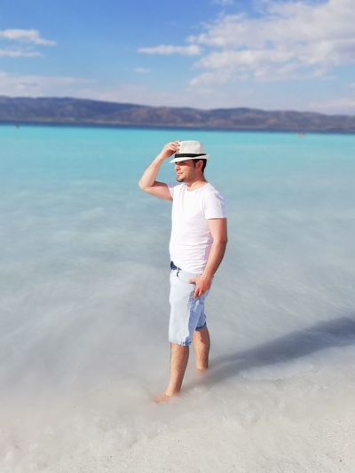 Full length of man holding hat while standing in sea