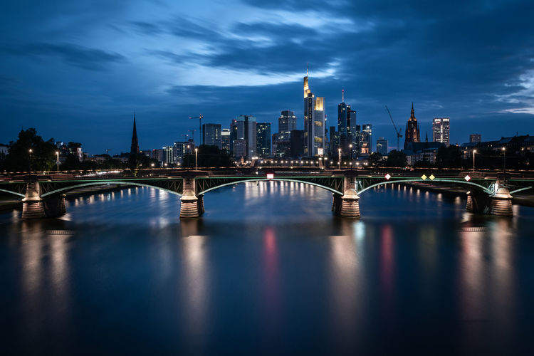 Illuminated bridge over river against cloudy sky at dusk