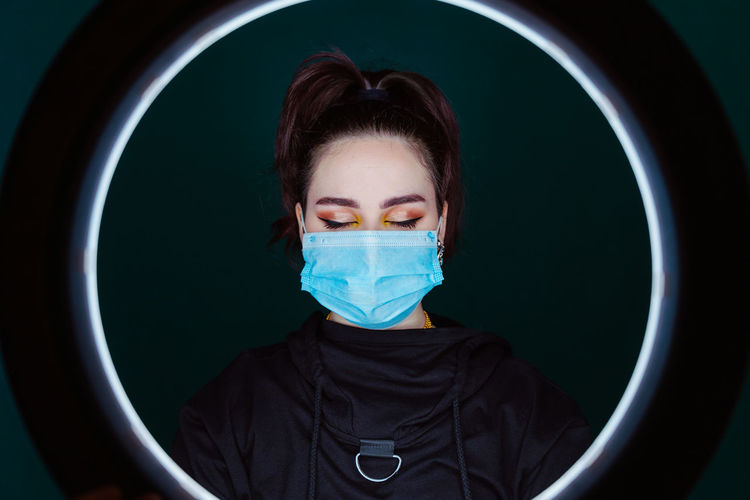 Calm female wearing medical mask standing in modern studio with eyes closed through ring lamp on black background