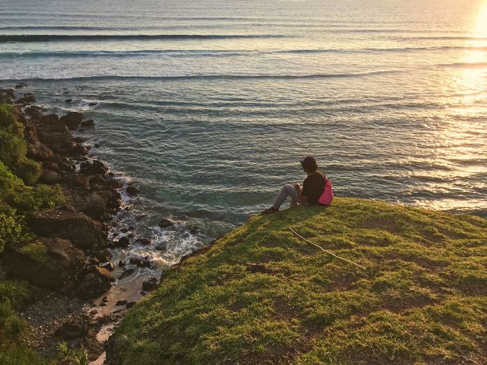 Man sitting on cliff against beach during sunset