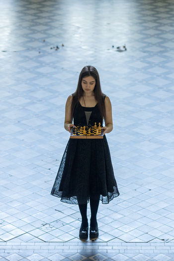 Beautiful girl in black dress plays chess in hall checkered floor