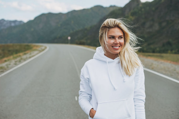 Beautiful smiling blonde young woman traveler in white hoodie on road, trip to mountains, altai