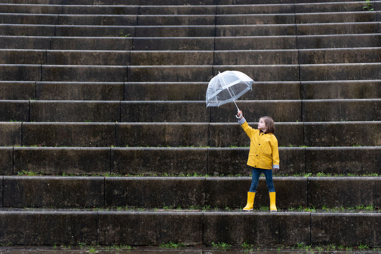 Kid in yellow raincoat and rubber boots playing under umbrella along stone steps on rainy day in city