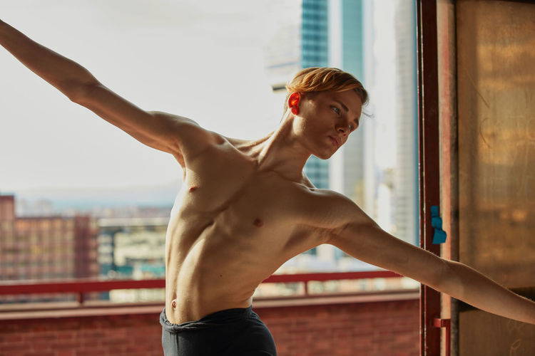 Amazing young gymnast in black leotard stretching the arm near window in room and looking away