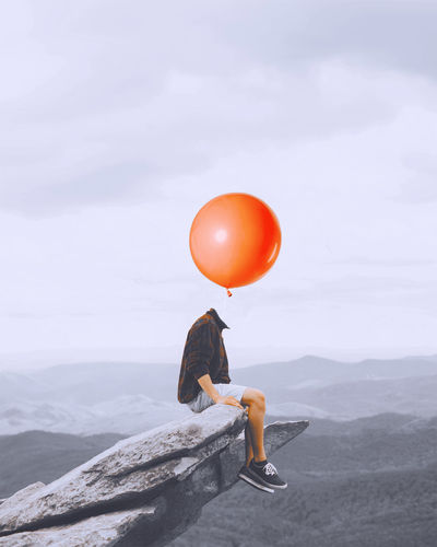 Digital composite image of decapitated man with balloon sitting on rock against sky