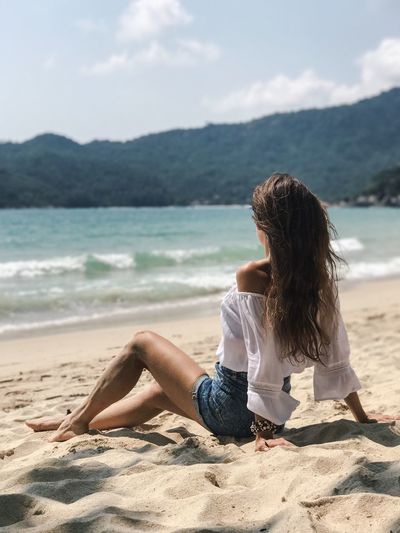 Side view of young woman sitting at sandy beach against mountains