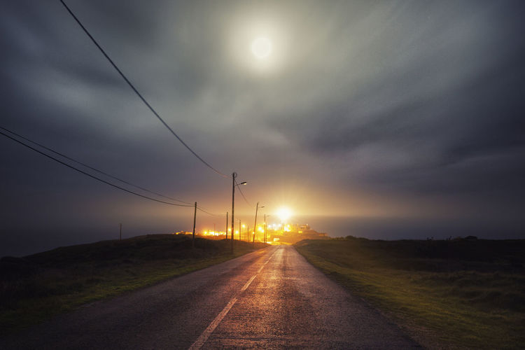 Diminishing perspective of empty road leading towards illuminated cabo da roca lighthouse against cloudy sky at night