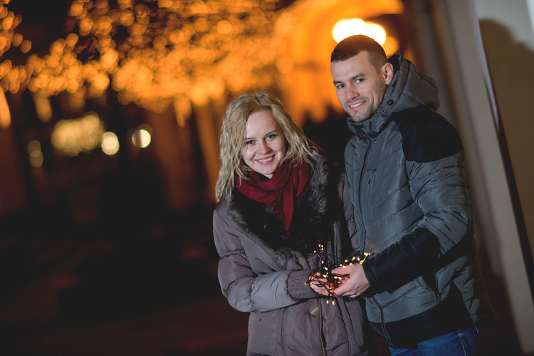 Smiling young couple standing while holding string lights during winter