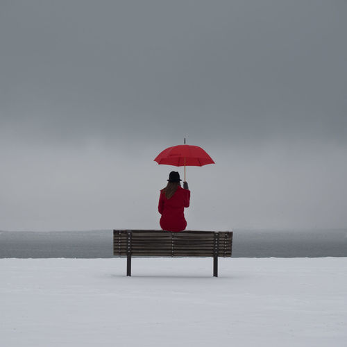 Woman with umbrella sitting on bench at snow covered field