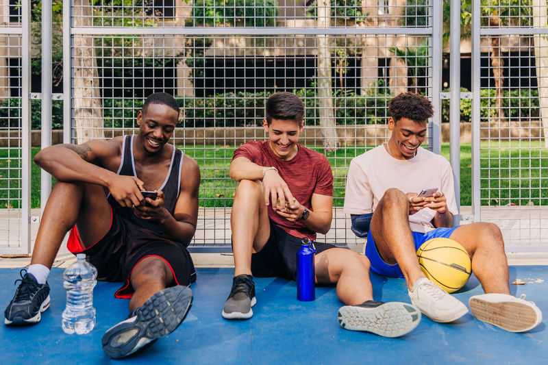 Cheerful young multiracial male athletes in sportswear browsing smartphones and laughing while sitting near net gate on sports ground