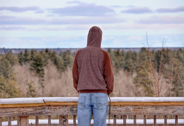 Rear view man wearing hood standing by railing while looking at forest against sky