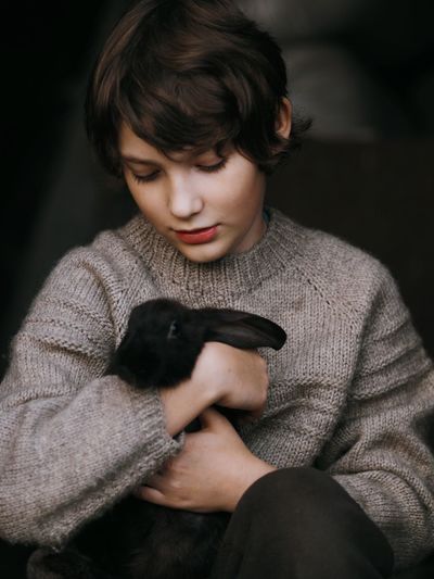 A cute teenage boy in a gray knitted sweater holds a small domestic black rabbit in his hands.