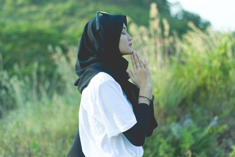 Side view of young muslim woman standing and pray on field