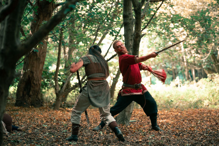 Full body of angry warrior in viking costume fighting opponent with sword during battle in forest