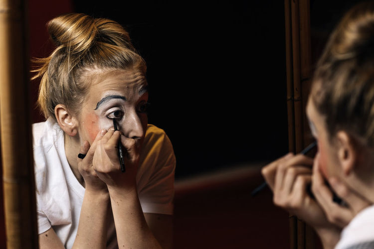 Young professional female mime in casual clothes painting the eyes and looking in mirror in dressing room