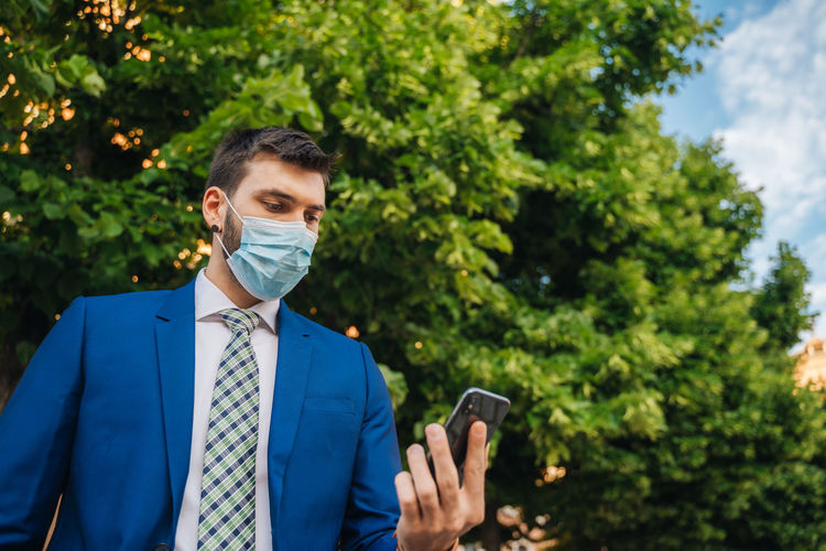 Young elegant businessman with a surgical mask looks at the phone in the park