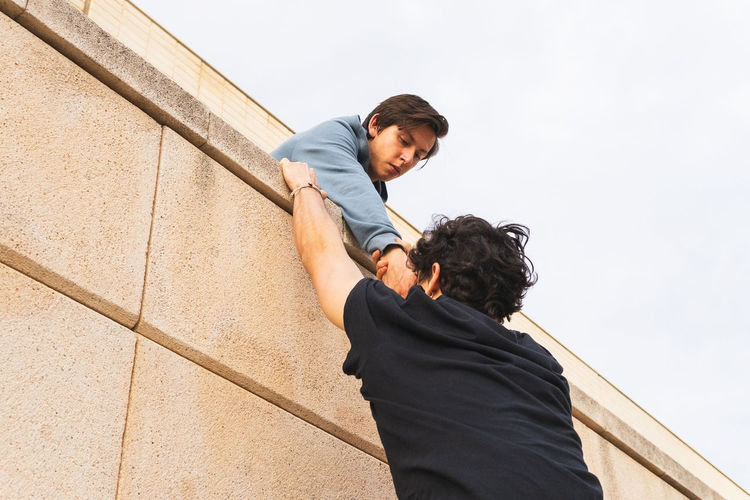 From below man in gray clothes on roof holding hand of man in strong clasp on outside