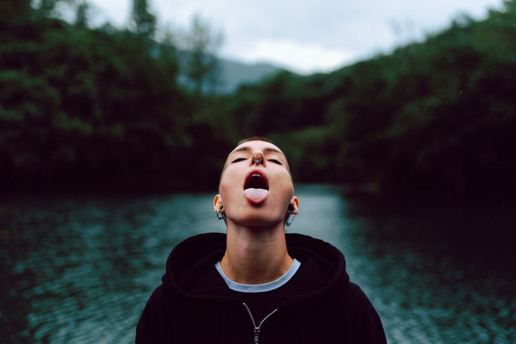 Short haired female with piercing wearing black hoodie looking up while catching raindrops with tongue near green forest and pond
