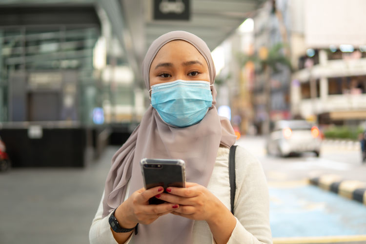 Portrait of young woman wearing mask holding phone while standing outdoors
