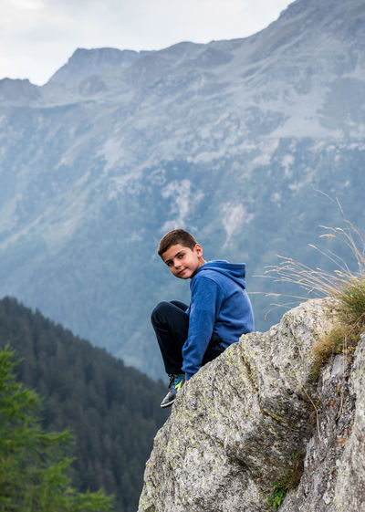 Boy sitting on rock formation against mountains