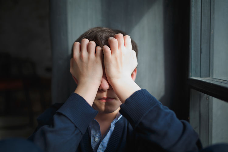 Pensive sad teenager boy with blue shirt and blue jeans sits at the window and closes his face with his hands. The sun's rays fall on the boy's face. Adult Body Part Boy Childhood Covering Depression - Sadness Embarrassment Emotion Finger Focus On Foreground Hand Hands Covering Eyes Headshot Hiding Human Body Part Human Face Human Hand Indoors Lifestyles Obscured Face One Person Portrait Real People Sadness Teenager