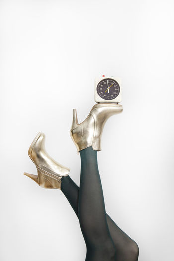 Woman wearing golden stilettos while balancing alarm clock against white background