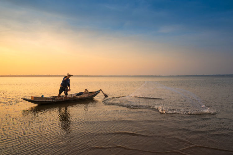 Fisherman casting net in lake against sky during sunset