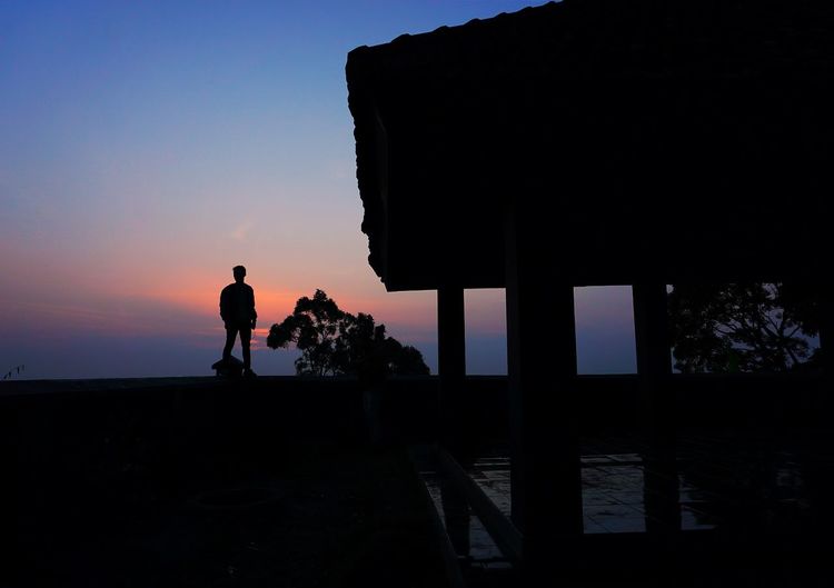 Silhouette man standing on retaining wall against sky during sunset