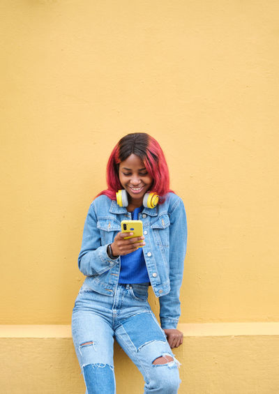 Happy young african american female hipster with pink dyed hair dressed in trendy denim outfit enjoying music through wireless headphones with smartphone while standing against yellow wall on street