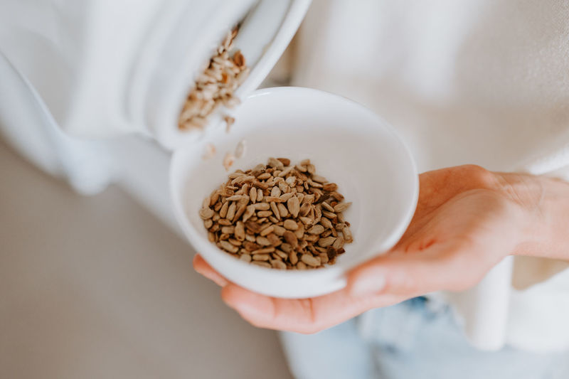 From above of crop unrecognizable female pouring sunflower seeds in bowl for healthy breakfast at home