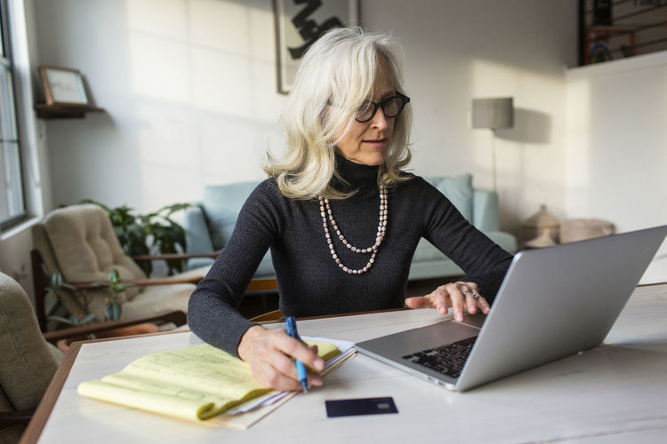Serious senior woman using laptop computer while paying bills at home