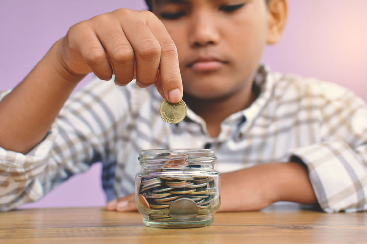 Close-up of boy putting coin in piggy bank at table against wall