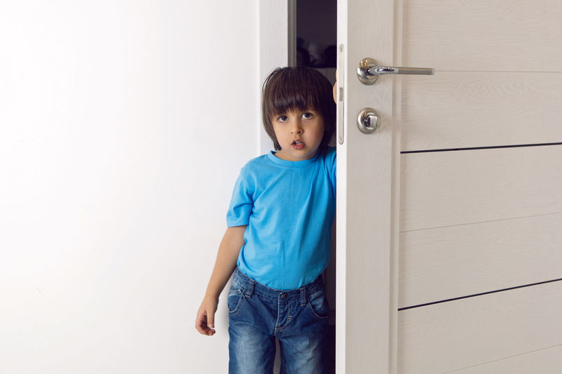 Boy in a blue t-shirt and jeans is standing at home in the white door