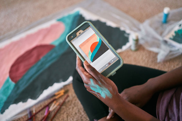 Crop unrecognizable african american female using app with picture sample on smartphone while learning painting at home during free time