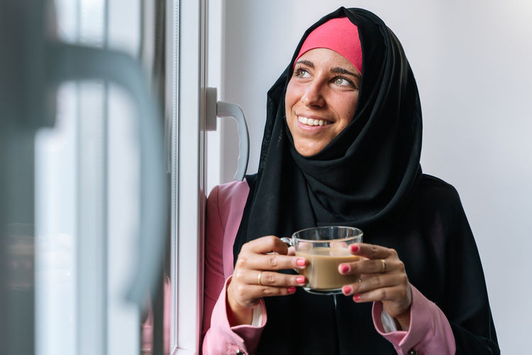 Smiling woman in hijab drinking coffee by window