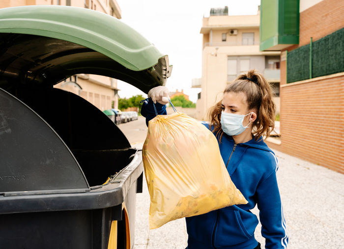Girl throwing the a yellow trash bag into a green recycling container open in the street. the teenager is wearing face mask and gloves to protect herself from infections, virus, bacterias. ho